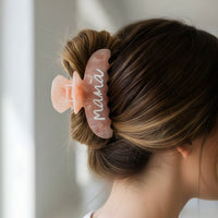 A brunette woman with a bun wears The Darling Effect’s Mama Word Up Claw Clip, a large pale pink clip with “mama” in white script. This bold accessory offers secure hold and style against an elegant, softly blurred background.