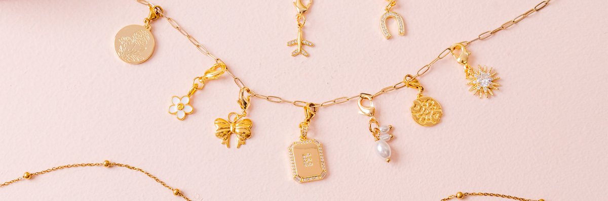 Gold charm necklaces and pendants are displayed on a light pink background. The charms include a bow, a pearl, a sunburst, flowers, and other decorative designs.