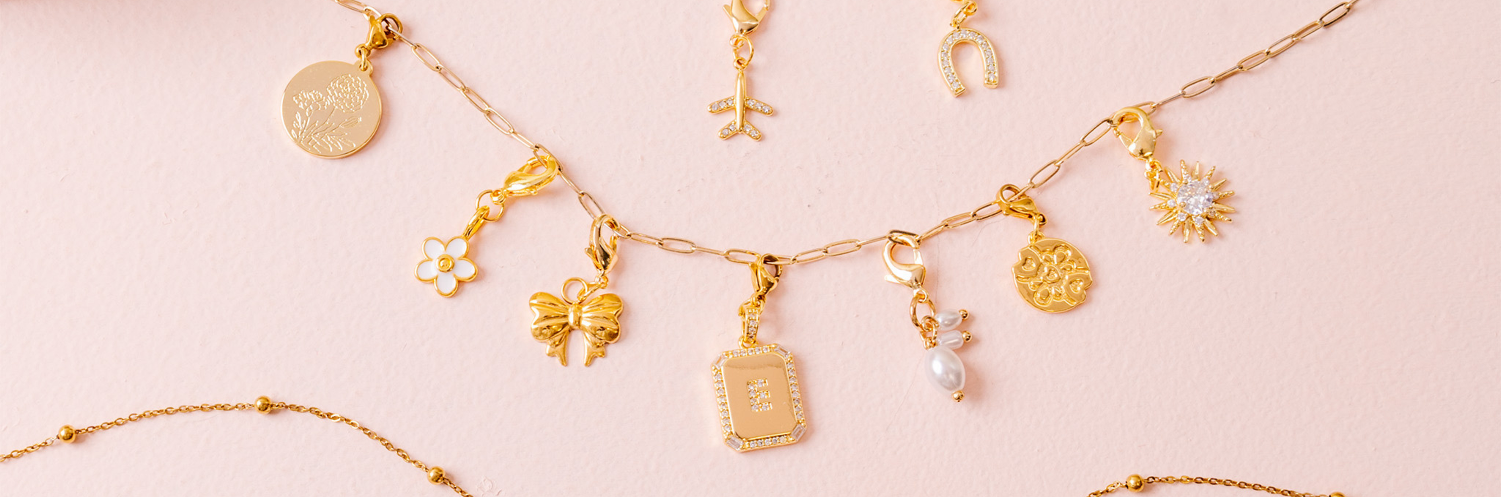 Gold charm necklaces and pendants are displayed on a light pink background. The charms include a bow, a pearl, a sunburst, flowers, and other decorative designs.
