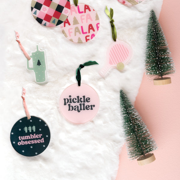 Holiday ornaments featuring playful designs and phrases, such as the “Holiday Tree Ornament - Pickleballer” by The Darling Effect, are displayed on white faux fur with two miniature bottlebrush pine trees against a pink and white backdrop. These festive Acrylic Holiday Ornaments capture the joyful spirit of the season perfectly.