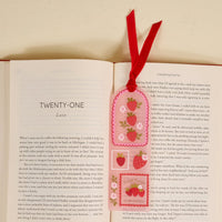 An open book shows chapter twenty-one. Resting on the right page is The Darling Effect's Farm Fresh Strawberries Acrylic Bookmark, adorned with strawberries, flowers, and a red ribbon. The background is a light cream surface—ideal for book lovers.