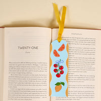 The Fruit Market Acrylic Bookmark, featuring orange slices, cherries, lemons, and a yellow ribbon, lies on the right page of an open book—ideal for book lovers or as a thoughtful gift.