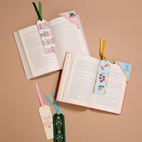 Three open books rest on a beige surface, each featuring The Darling Effect’s Fruit Market Acrylic Bookmark—a colorful accessory with fruit and floral designs. Two more bookmarks are placed nearby, creating a charming gift for book lovers.