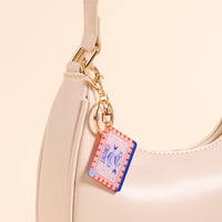 Close-up of The Darling Effect’s Sardines Acrylic Keychain, featuring pink and red stripes and fish illustrations, attached to a cream handbag with a gold clasp. The beige background enhances this stylish accessory's charm.