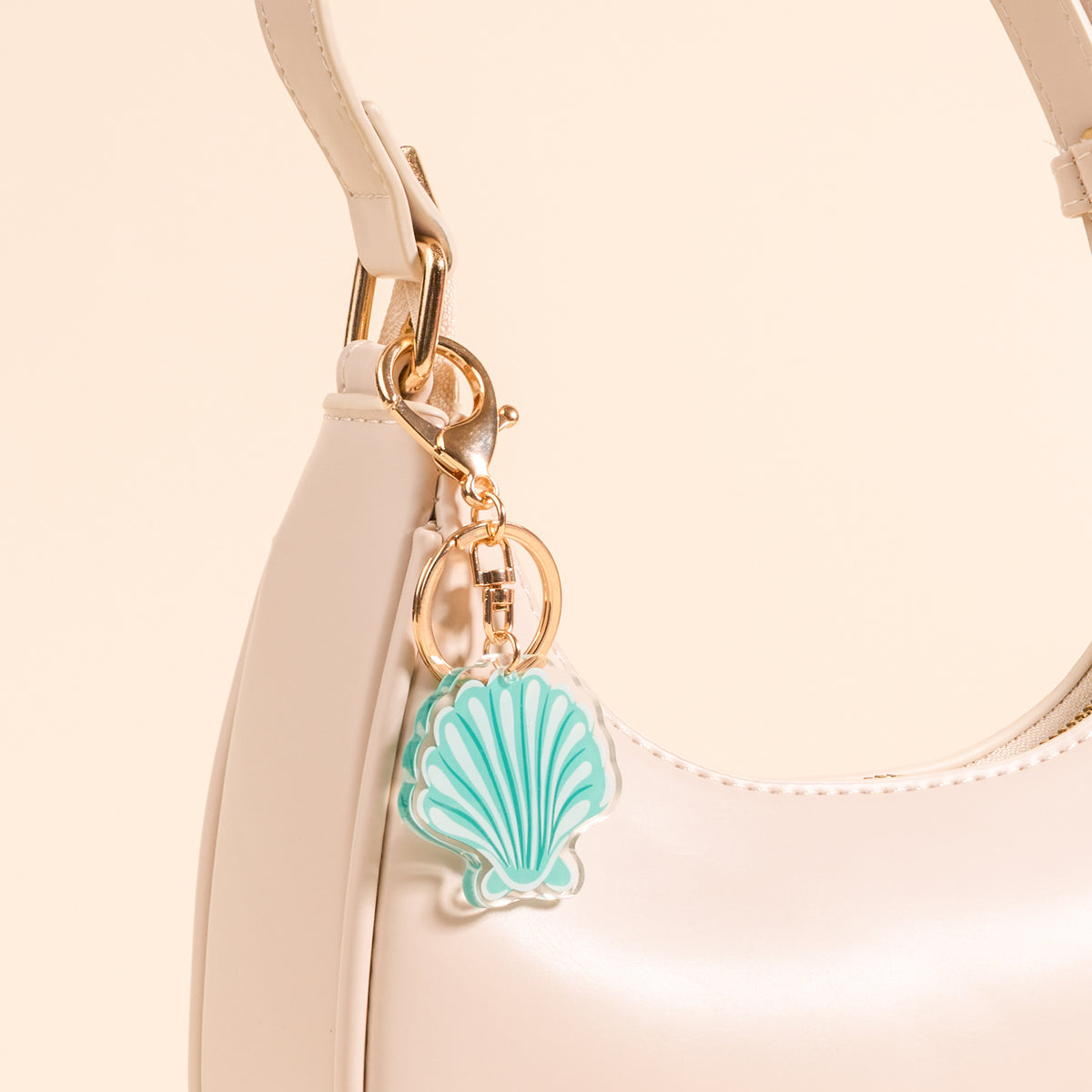 A cream-colored handbag with gold hardware features The Darling Effect’s Seashell Acrylic Keychain—a cute turquoise and white seashell-shaped accessory—attached to one of the straps.