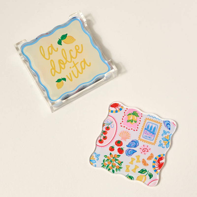 The Darling Effect’s Ciao Bella Table Talk Acrylic Coaster Set features two decorative scalloped-edge coasters—one in a clear holder with “la dolce vita” and lemons, one with colorful Italian-inspired art—perfect for any table.