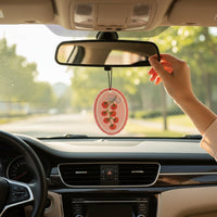 A hand hangs The Darling Effect’s Off the Vine Sweet Ride Car Air Freshener, featuring a cherry tomato vine design, on the rearview mirror with a sunny, tree-lined road visible through the windshield.