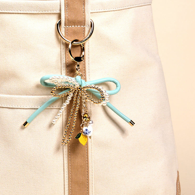 Close-up of a beige canvas bag with tan straps, featuring The Darling Effect's Limoncello Love Layered Bows Bag Charm—adorned with a mint green bow, pearls, gold chains, a floral bead, and a yellow charm on a ring.