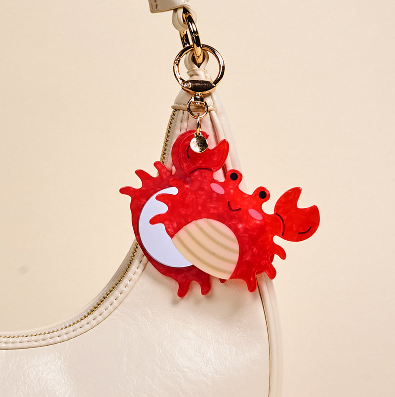 The Darling Effect's Crab Mini Mirror Bag Charm, shaped like a cute red crab, hangs from the handle of a white handbag against a plain beige background.