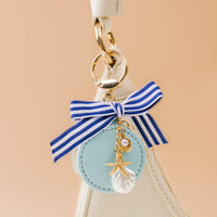 A close-up of The Darling Effect's Sea Breeze Travel Jewelry Case Bag Charm with a blue and white striped bow, gold starfish, pearl, seashell, and light blue charm—perfect for accessorizing your portable jewelry organizer.