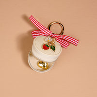 The Darling Effect Strawberry Shortcake Travel Jewelry Case Bag Charm is a small, cream round organizer with a gold keyring, gingham bow, and strawberry charms, holding a shiny gold heart pendant. Background: light beige surface.