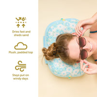 A woman in sunglasses relaxes on The Darling Effect’s Daisy Craze Aqua Ballast Beach Pillow. Icons highlight its fast-drying, sand-shedding plush top and wind-resistant design. Shop this limited-edition collab now!.