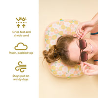 A woman relaxes on The Darling Effect’s Daisy Craze Peach Ballast Beach Pillow, smiling as icons highlight: “Dries fast and sheds sand,” “Plush, padded top,” and “Stays put on windy days.”.