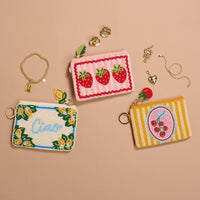 Three Ciao Handcrafted Beaded Pouches by The Darling Effect—featuring strawberries, "Ciao" lemons, and cherries—are displayed with gold jewelry on a tan background.
