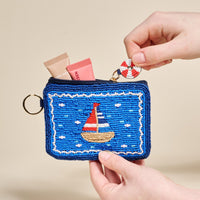 The Darling Effect's Sail Away Handcrafted Beaded Pouch in blue, held by two hands, features a sailboat design and comes with a small life preserver keychain attached to the zipper.