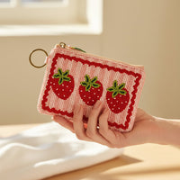 A hand holds The Darling Effect’s Strawberries Handcrafted Beaded Pouch, adorned with three red strawberries and green leaves on a pink and white base, featuring a gold keyring—all shown in soft, natural light.