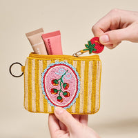 A hand holds The Darling Effect’s Off the Vine Handcrafted Beaded Pouch, a compact yellow accessory adorned with a charming cherry design and two cosmetic tubes inside. The zipper pull is accented by an adorable beaded strawberry, making this pouch the perfect way to carry your essentials in style.