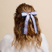 A person with wavy, light brown hair styled half-up and tied with The Darling Effect’s All-American Darling Lovely Layered Hair Bow is shown from behind in a white textured top against a beige background, highlighting their chic taste in accessories.