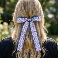 Wearing The Darling Effect's All-American Darling Lovely Layered Hair Bow, a person with long, wavy blonde hair stands outdoors with their back to the camera. The bold blue, white, and red accessory makes an eye-catching statement.