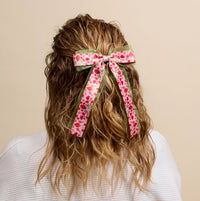 Seen from behind, a person with wavy, shoulder-length hair wears The Darling Effect's Strawberry Fields Lovely Layered Hair Bow—a large pink and green floral accessory—paired with a white textured top against a beige background.