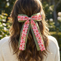 A person with wavy brown hair wears The Darling Effect's Strawberry Fields Lovely Layered Hair Bow, a large pink floral accessory with green edging, tied at the back of their head while outdoors in a cream sweater.
