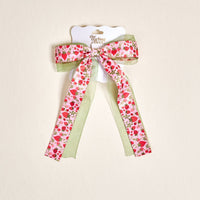 The Darling Effect's Strawberry Fields Lovely Layered Hair Bow features a pink bow with a strawberry pattern over a light green ribbon, attached to a branded card—perfect for fans of cute hair accessories.