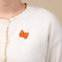 A person wears a white knit cardigan with large buttons, adorned on the left chest with The Darling Effect’s Cutie Crab Timeless Brooch—a charming enamel pin shaped like an orange crab.
