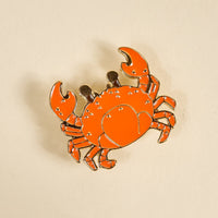 The Darling Effect's Cutie Crab Timeless Brooch features an orange enamel crab with raised claws and black eyes, outlined in gold against a beige background—a vintage-inspired keepsake that adds charm to any collection.