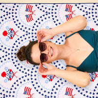 A smiling woman in sunglasses relaxes on The Darling Effect’s All-American Darling XL Quick-Dry Beach Towel, which features a patriotic design with small American flags, stars, and red, white, and blue patterns. She wears a teal top and a necklace.