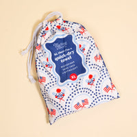 A drawstring bag with a patriotic pattern featuring American flags and eagles, labeled “hi, there! I’m the All-American Darling XL Quick-Dry Beach Towel by The Darling Effect” in blue and white, is shown on a light beige background. Made from soft microfiber fabric, this sand-resistant towel is perfect for your next beach adventure.