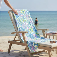 A Coastal Cove XL Quick-Dry Beach Towel by the-darling-effect, featuring a colorful sea turtle and flower design, covers a beach chair on a patio with an ocean view. In the background, a woman in a swimsuit walks into the water.