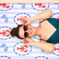 A smiling woman in sunglasses and a teal swimsuit relaxes on the Harbor House XL Quick-Dry Beach Towel by The Darling Effect, featuring red lobsters and blue seashells, looking up at the camera.