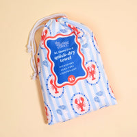 The Darling Effect Harbor House XL Quick-Dry Beach Towel comes in a blue and white striped drawstring bag with red lobsters. It's microfiber, sand-free, lightweight—perfect for all your adventures.