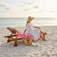 A woman in a white dress and straw hat sits on a lounge chair with The Darling Effect’s Poppin' Petals Hot Pink XL Quick-Dry Beach Towel, facing the ocean as the sun sets. A straw bag and open book lie on the sand beside her.