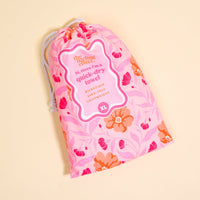 A pink drawstring bag with a floral pattern displays a label reading, “The Darling Effect Poppin' Petals Hot Pink XL Quick-Dry Beach Towel, microfiber, sand-free, lightweight,” on a pale yellow background.