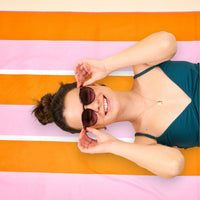 A smiling woman in sunglasses relaxes on The Darling Effect Sicily Stripes Pink XL Quick-Dry Beach Towel.
