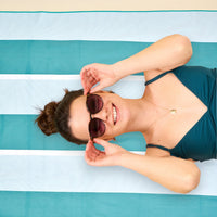 A woman in a teal swimsuit relaxes on The Darling Effect’s Sicily Stripes Teal XL Quick-Dry Beach Towel, smiling and adjusting her sunglasses with her hair in a bun.