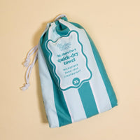 A teal and light blue striped drawstring bag labeled "The Darling Effect" sits on a beige background, containing the Sicily Stripes Teal XL Quick-Dry Beach Towel—microfiber, sand-free, lightweight, and perfect for the beach.