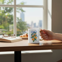 A hand holds The Darling Effect's Capri Citrus 11 oz Cozy Cute Ceramic Mug, featuring a golden handle and vibrant lemon-leaf design, at a wooden table beside an open book, eyeglasses, and a croissant in a sunlit city-view room.