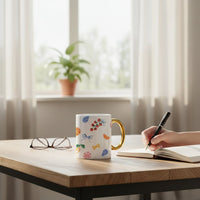 A person writes in a notebook at a wooden table with The Darling Effect’s Italian Summer 11 oz Cozy Cute Ceramic Mug, eyeglasses, and a potted plant on the windowsill. Sunlight pours through sheer curtains, adding warmth to the cozy scene.