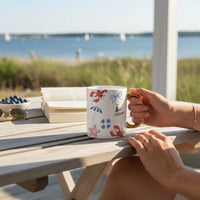 Enjoy coffee or tea at a wooden table outdoors with The Darling Effect's Seaside Haven 11 oz Cozy Cute Ceramic Mug, featuring colorful beach-themed designs and a golden handle. Nearby: an open book, sunglasses, blueberries, and views of sailboats.