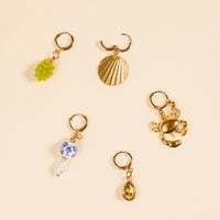 A flat lay of five gold-toned hoop earrings from The Darling Effect’s Amalfi Coast The Ultimate Charm Pack, each featuring a unique charm: green beaded grapes, ribbed shell, scorpion, porcelain bead with pearls, and citrine gemstone.