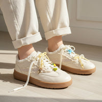 A person wears cream-colored sneakers with The Darling Effect's Amalfi Coast The Ultimate Charm Pack on the laces, paired with light-cuffed pants, standing indoors on a light wood floor.