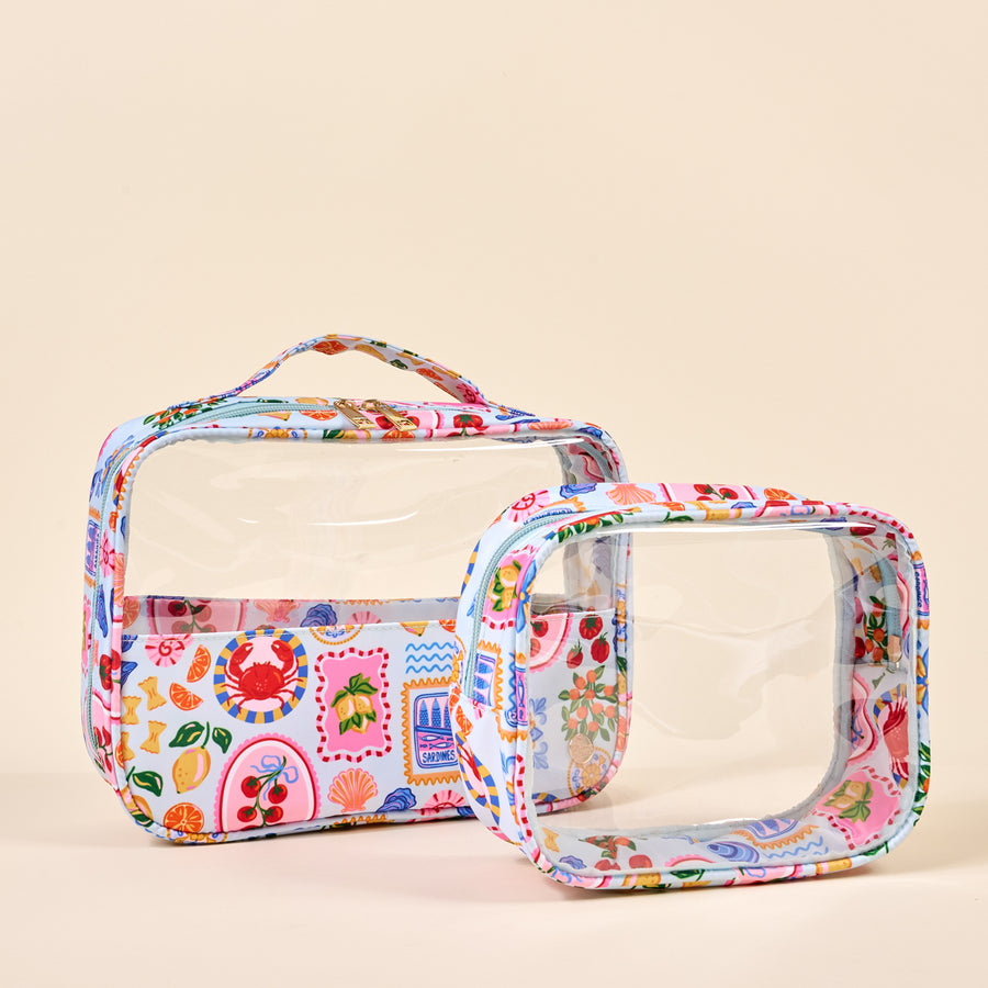 The Darling Effect's Ciao Bella Clearly Chic Cosmetic Bag Set features two clear bags with playful, patterned trims—a roomy handled toiletry bag and a matching smaller bag, both set against a chic beige background.