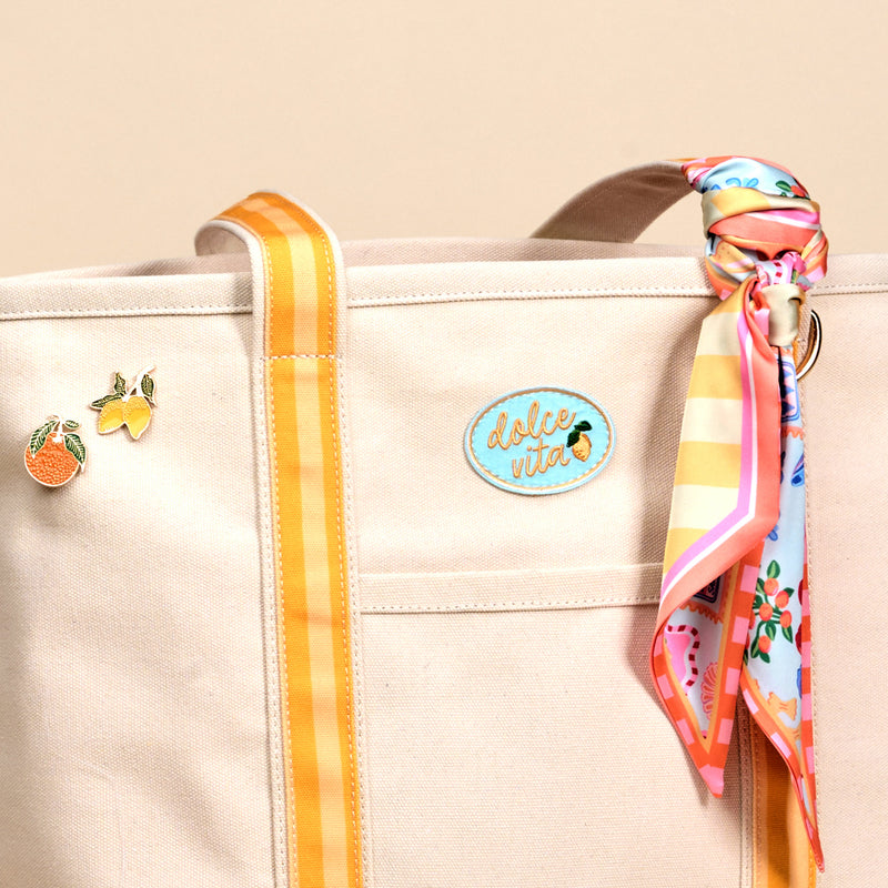 The Darling Effect’s Ciao Bella The Darling Canvas Tote Bag features comfy straps, yellow striped handles, fruit pins, a “dolce vita” patch, and a colorful scarf accent. Its spacious interior holds all your essentials.
