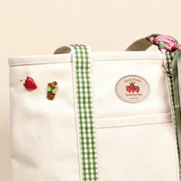 The Darling Effect’s Strawberry Fields The Darling Canvas Tote Bag features light canvas, comfy straps, green gingham trim, a strawberry pin, strawberry plant pin, Farm Fresh Strawberries patch, and a green and pink strawberry-patterned scarf.