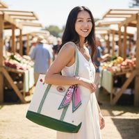 A woman in a sleeveless white dress smiles while holding The Darling Effect’s Strawberry Fields The Darling Canvas Tote Bag, its spacious interior ideal for farmers market finds. A patterned scarf is attached, adding flair to the green-accented tote.