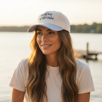 A woman with long brown hair smiles in casual style, wearing The Darling Effect's Meet Me at the Lake Classic Canvas Hat against a softly blurred lake sunset. The beige hat features "meet me at the lake" embroidery.