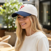 A smiling woman with long blonde hair wears The Darling Effect's Lobster Classic Canvas Hat—white and navy with a red lobster design—paired with a relaxed fit white shirt as she sits outdoors at a cafe surrounded by plants.