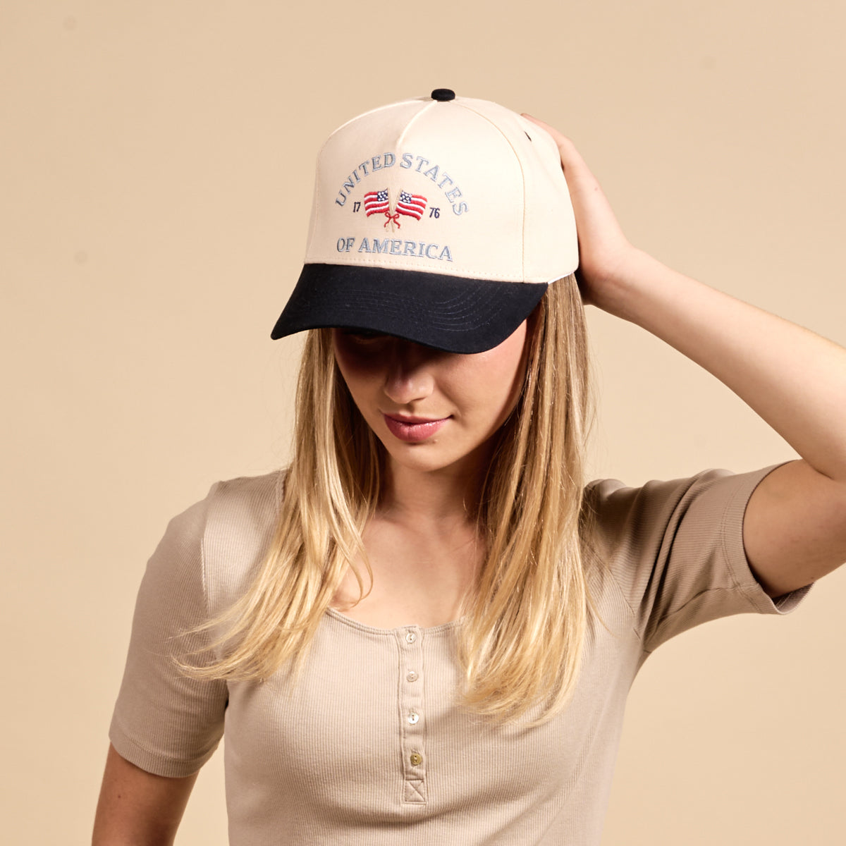 A woman sports The Darling Effect's United States of America Classic Canvas Hat—cream and navy with "1776" and crossed flag embroidery—paired with a beige ribbed henley for an easy, laid-back style.